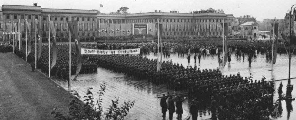 Niemieckie uroczystości z okazji pierwszej rocznicy wybuchu II wojny światowej połączone z przemianowaniem placu Piłsudskiego na Adolf Hitler Platz. Na placu liczne wojska niemieckie, flagi ze swastyką oraz transparent z napisem: "Adolf Hitler ist Deutschland", pogoda deszczowa.