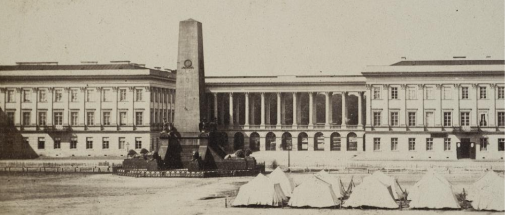 Pałac Saski od strony placu Piłsudskiego, na pierwszym planie pomnik w formie obelisku, przed nim po prawej stronie dziesięć namiotów wojskowych ustawionych w dwóch rzędach.