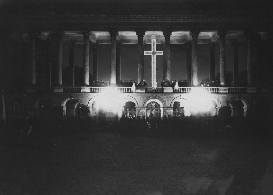 Podświetlona kolumnada Pałacu Saskiego wyłaniająca się z ciemności nocy. Po środku podświetlony jest Grób Nieznanego Żołnierza, a nad nim świecący krzyż.