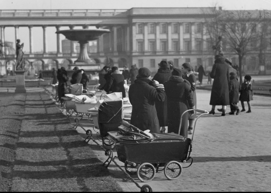 Rząd wózków dziecięcych wraz z opiekunkami stojącymi w alejce Ogrodu Saskiego. W tle kolumnada i jedno skrzydło Pałacu Saskiego.