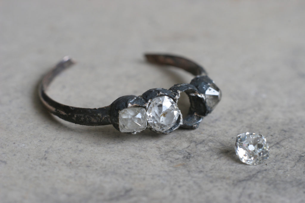 Pierścionek ze złota i srebra, pokryty czarnym nalotem. Ozdobiony czterema diamentami, jeden poza oczkiem, obok pierścionka.