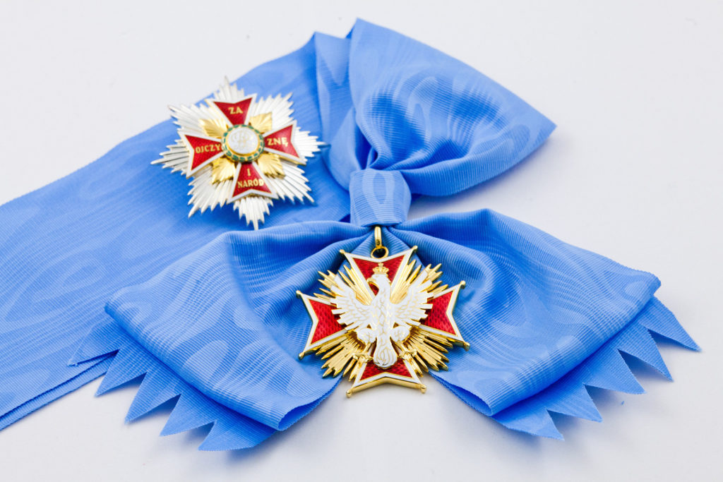 Order Orła Białego położony na ozdobnej wstędze. Na medalu znajduje się orzeł na tle krzyża równoramiennego.