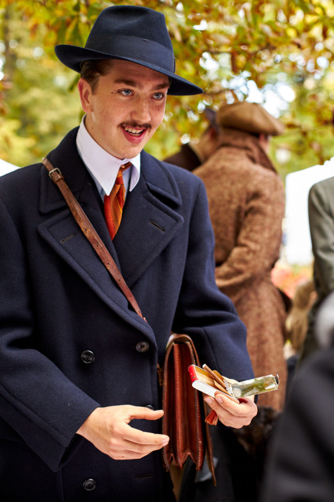 Uśmiechnięty animator wydarzenia ubrany w płaszcz, kapelusz i białą koszulę z krawatem trzyma w ręku paczkę cygaretek. Łokciem przytrzymuje starą skórzaną aktówkę koloru brązowego.