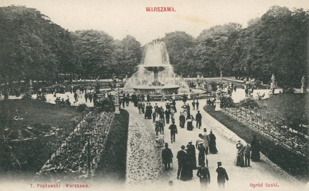 Archiwalne zdjęcie przedstawia klimat przedwojennej Warszawy. Duża grupa ludzi spaceruje główną aleją Ogrodu Saskiego w stronę tryskającej wodą fontanny oraz wokół niej.