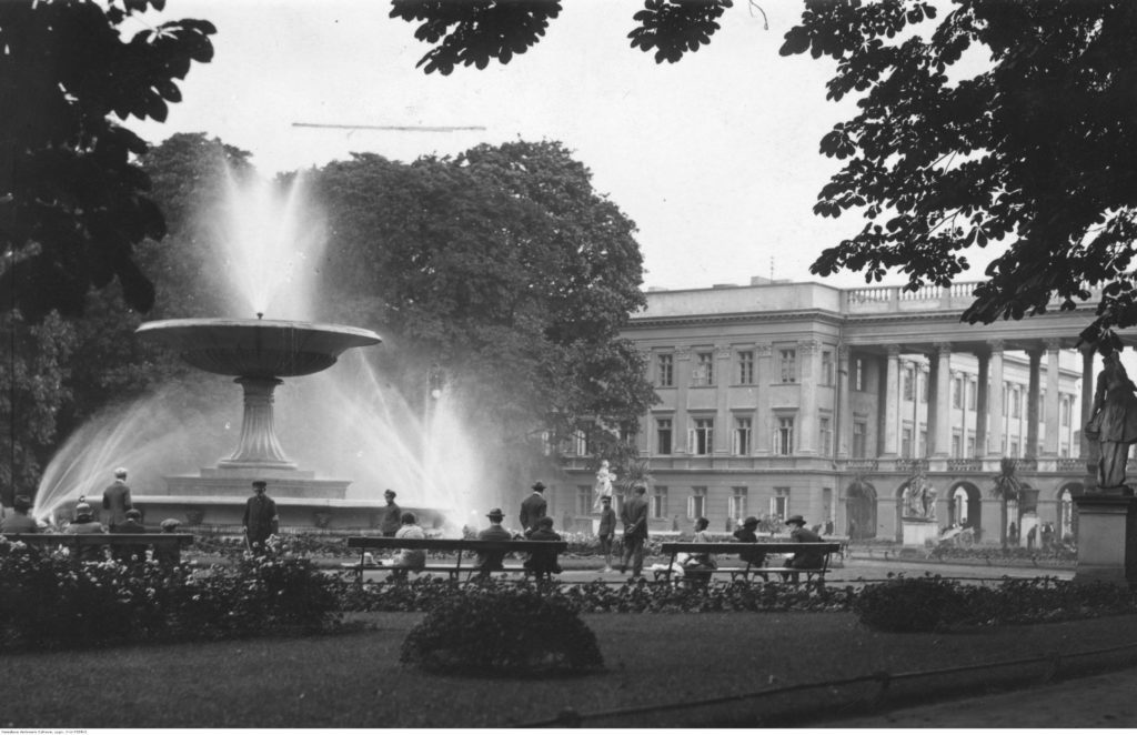 Archiwalne zdjęcie przedstawiające dużą fontannę w parku, wokół której siedzą ludzie na ławkach. Obok, pomiędzy drzewami wyłania się fragment gmachu Pałacu Saskiego z kolumnadą.