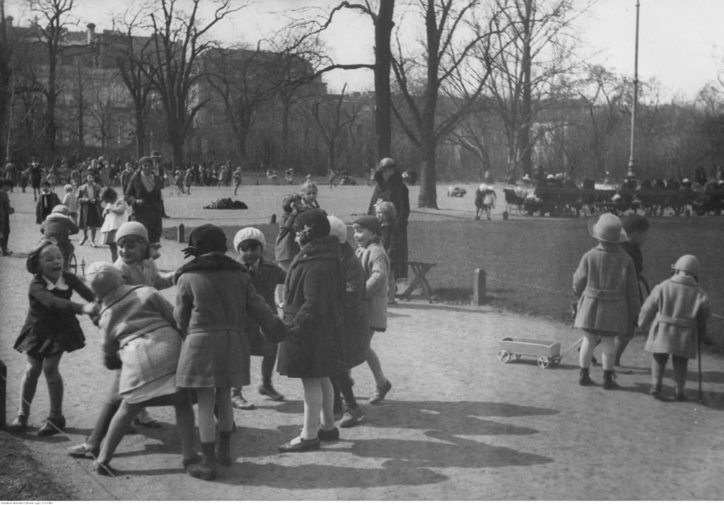 Archiwalne zdjęcie dzieci bawiących się razem na jednej z alejek Ogrodu Saskiego. Dzieci mają na sobie płaszczyki i berety, w tle wyłaniają się nagie drzewa.
