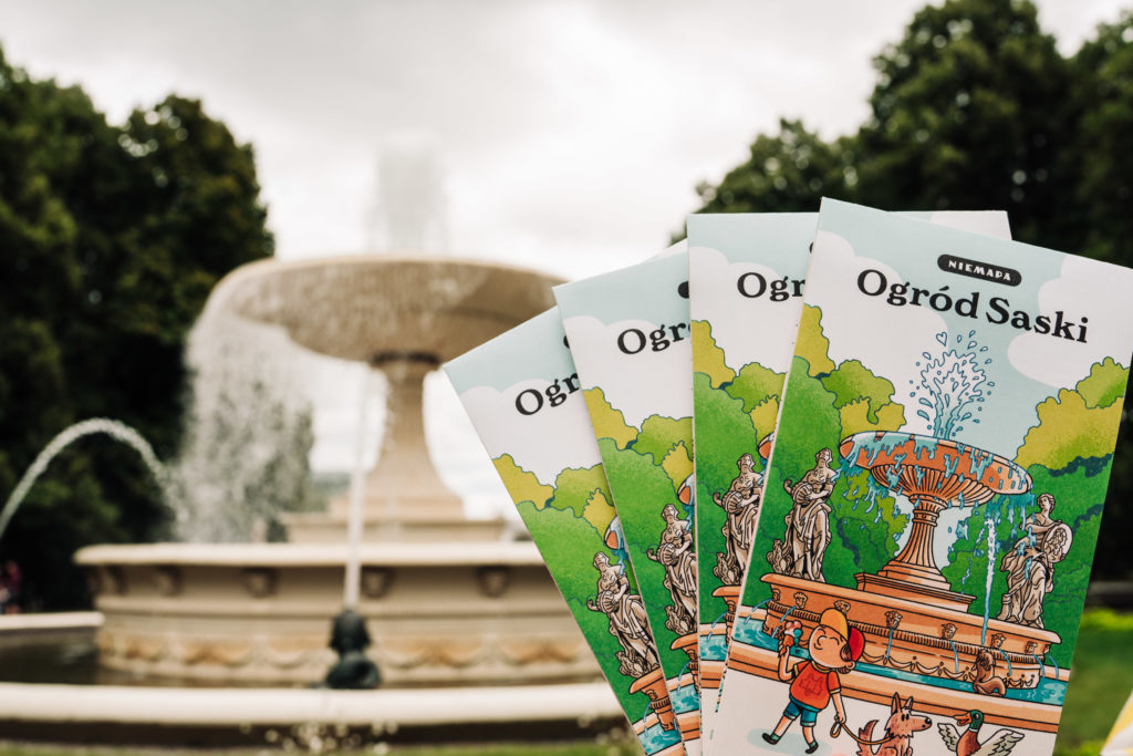 Four colorful folded maps held like a fan, in front of a large garden fountain.