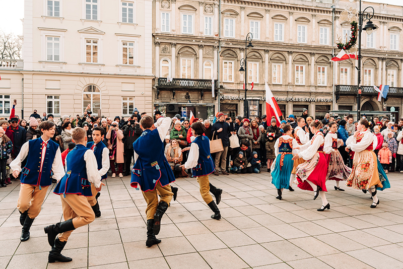 Na chodniku Krakowskiego Przedmieścia młodzież ubrana w polskie stroje ludowe tańczy w dwóch kręgach podczas Festiwalu Niepodległa na Krakowskim Przedmieściu. Dookoła tłumy gapiów oglądają ich występ.