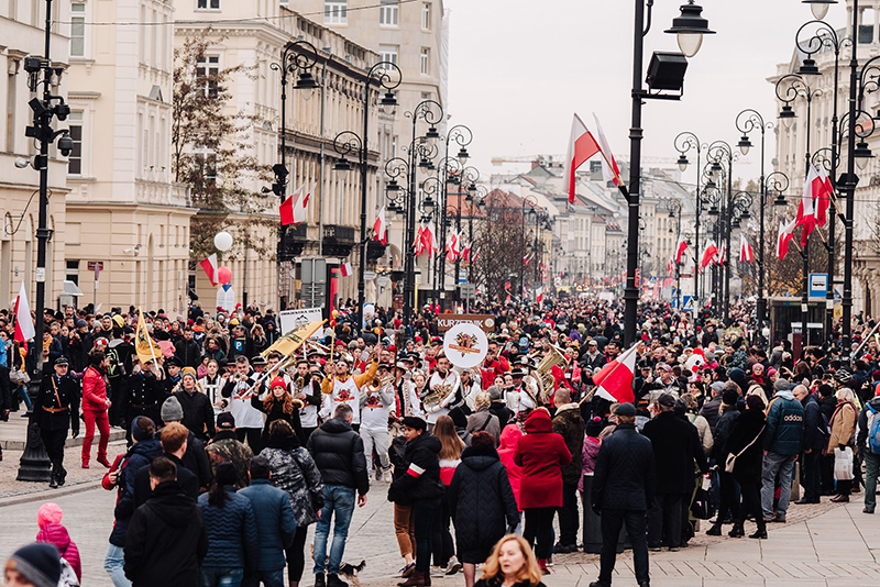 Tłumy na chodnikach śledzą przemarsz orkiestr, które idą środkiem ulicy podczas Festiwalu Niepodległa na Krakowskim Przedmieściu. 