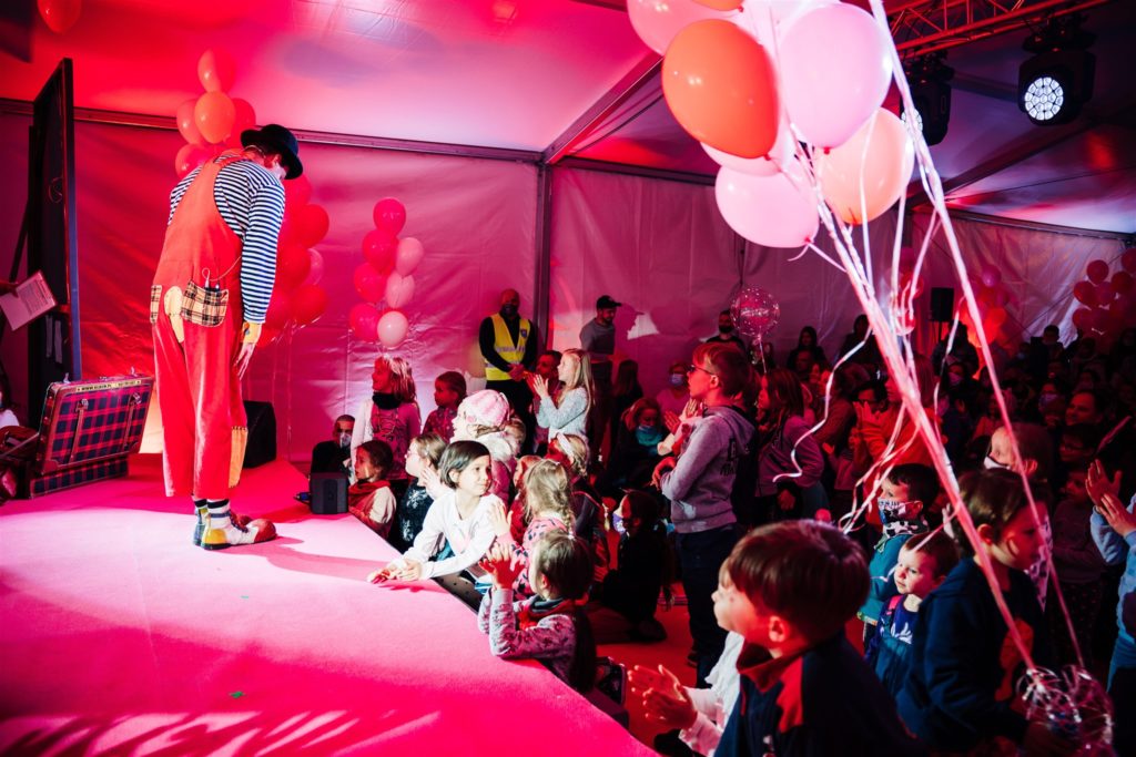 Klaun na scenie pod dużym namiotem rozmawia z dziećmi, które stoją poniżej sceny. Na pierwszym planie baloniki.