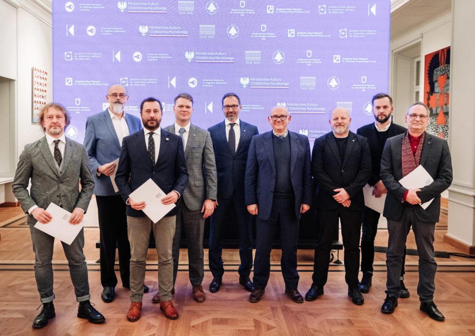 A photo of nine men in formal clothing. They are standing in front of a wall covered with logos of art academies and of the Pałac Saski company.