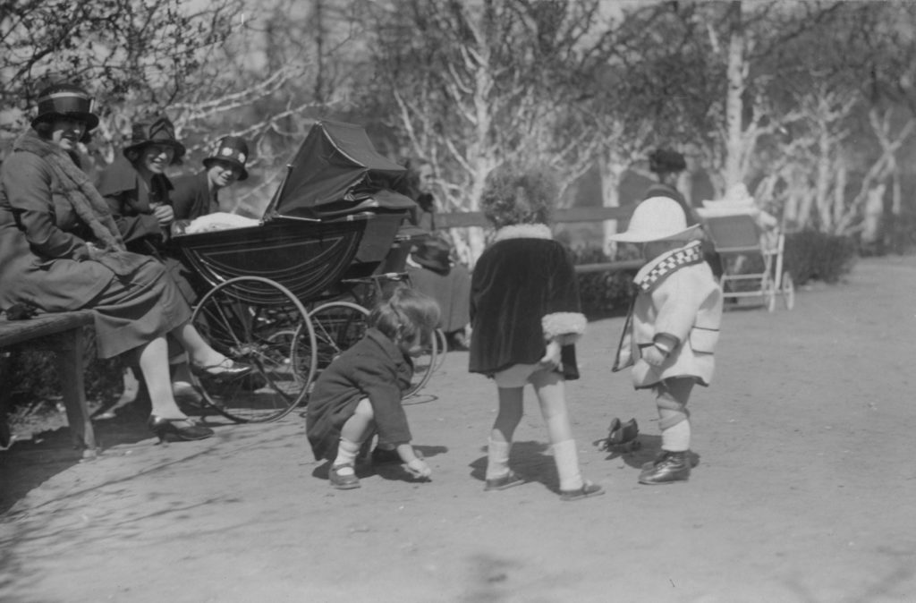Czarno-białe zdjęcie przedstawia grupę dzieci bawiących się w alejce Ogrodu Saskiego. Po lewej stronie na ławce siedzą uśmiechnięte niańki w strojach z lat 20. XX wieku.