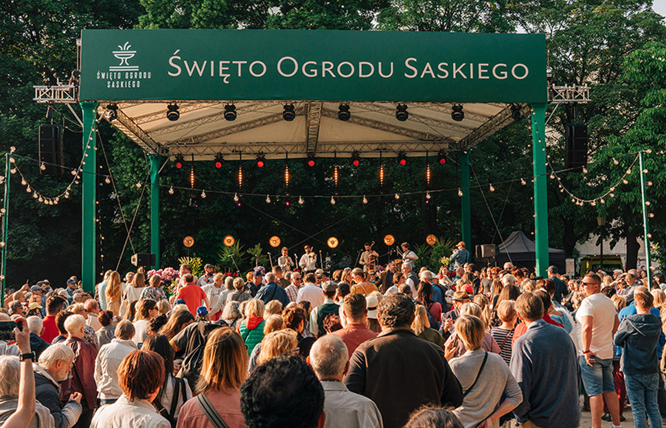 A view of the musical stage during the Saxon Garden Festival. There is a band playing on the stage, with people dancing and listening in front of the stage. There are tall trees in the background.