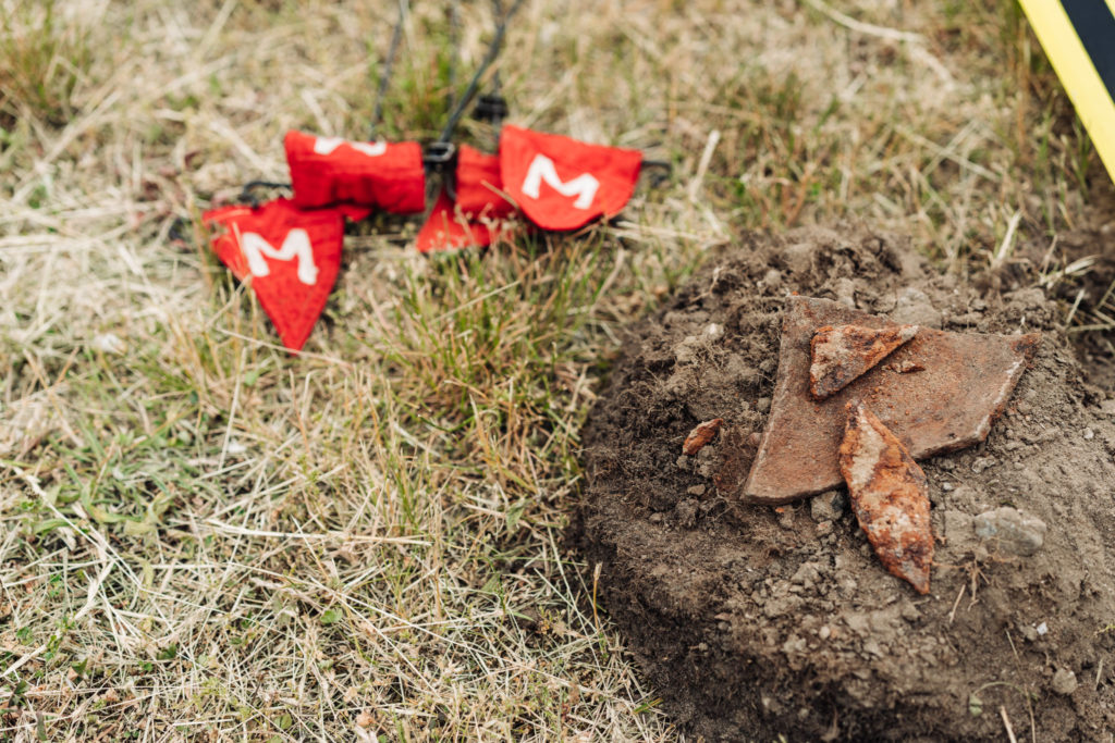 A detail of the first metal fragments dug up during the sapper survey which proceeds the archaeological works at the Brühl Palace area. Next to the couple of metal shards on a newly turned soil, there is a couple of small flags lying on the grass. The flags are used to mark locations of found objects.