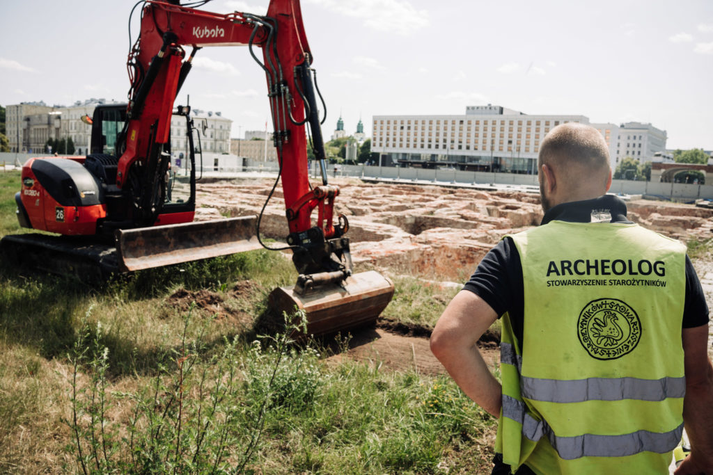 Prace archeologiczne na terenie Pałacu Brühla. Na pierwszym planie archeolog w żółtej kamizelce z napisem "ARCHEOLOG STOWARZYSZENIE STAROŻYTNIKÓW" nadzorujący pracę koparki ściągającej z ziemi warstwę trawy. W tle odsłonięte już relikty Pałacu Saskiego.