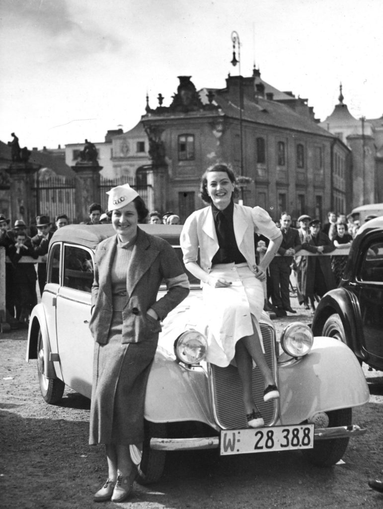 Jedna kobieta oparta o karoserię samochodu, druga siedzi na jego masce przed startem w rajdzie samochodowym kobiet, w tle należący w międzywojniu do MSZ Pałac Bruhla, międzywojnie.