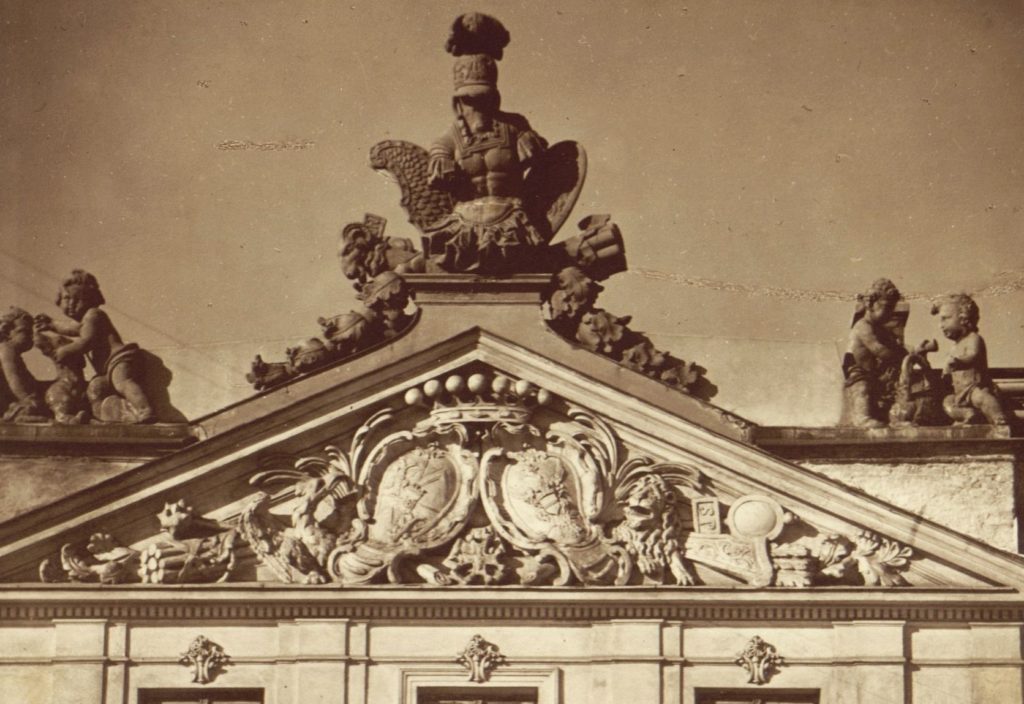An archive photo of a decorative, triangular surface at the top of a building. The triangle is filled with two coats of arms surrounded by small sculptures. On top of the triangular tympanum there is a sculpture of an armored torso with a helmet.