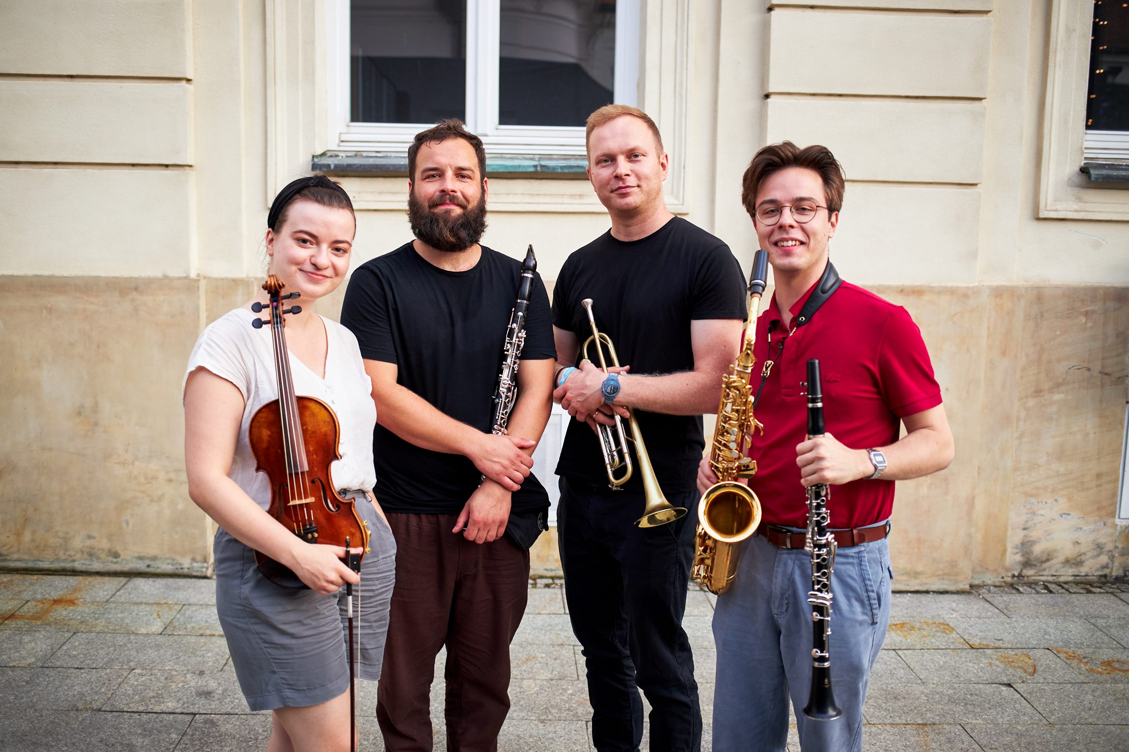 Czterech muzyków pozuje na tle budynku z instrumentami. Mają w rękach skrzypce, klarnet, trąbkę i saksofon.