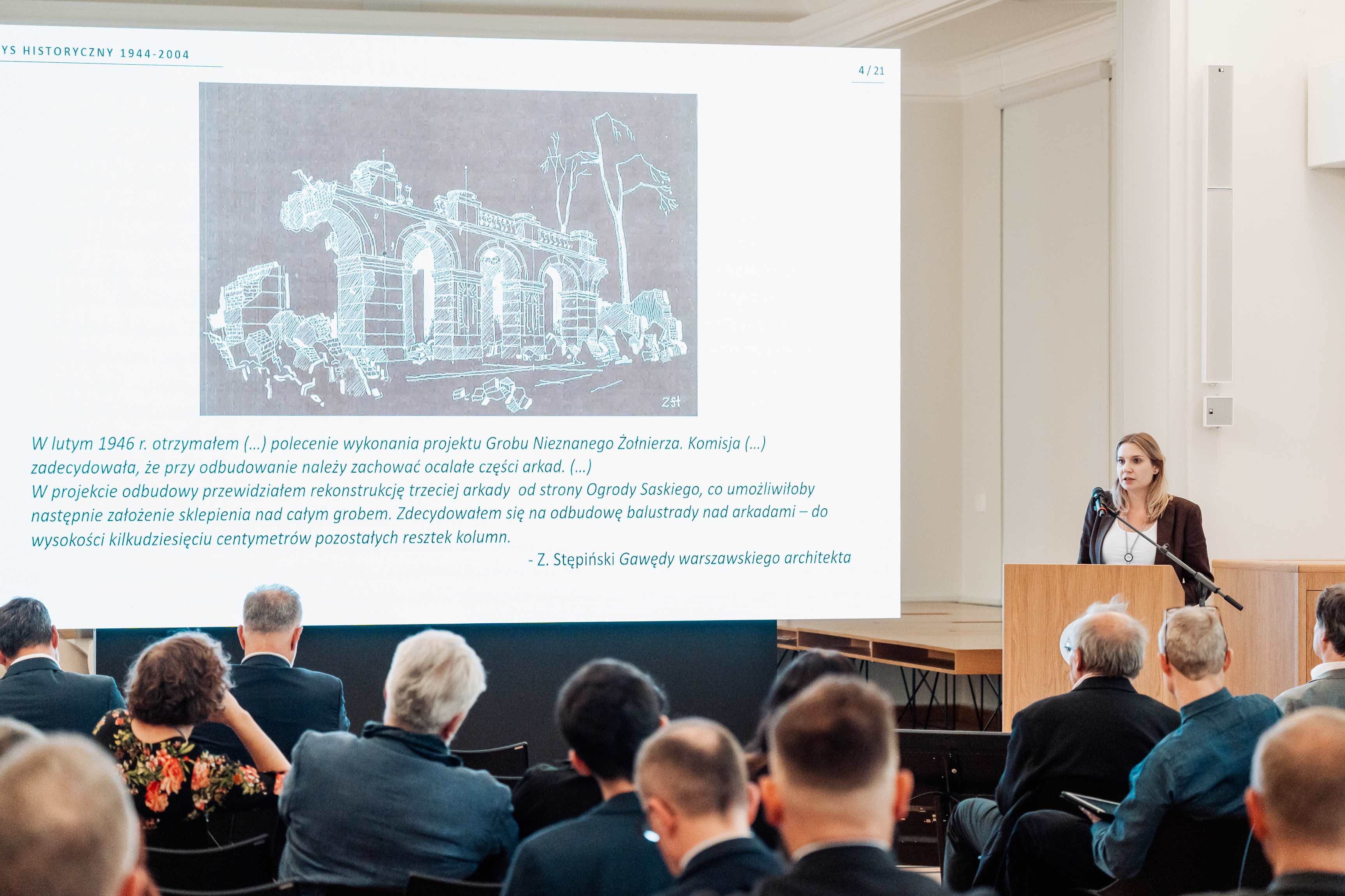 Maria Wardzyńska prezentuje prezentację na temat odbudowy Pałacu Saskiego. W tle ekran ze zniszczeniami Grobu Nieznanego Żołnierza.