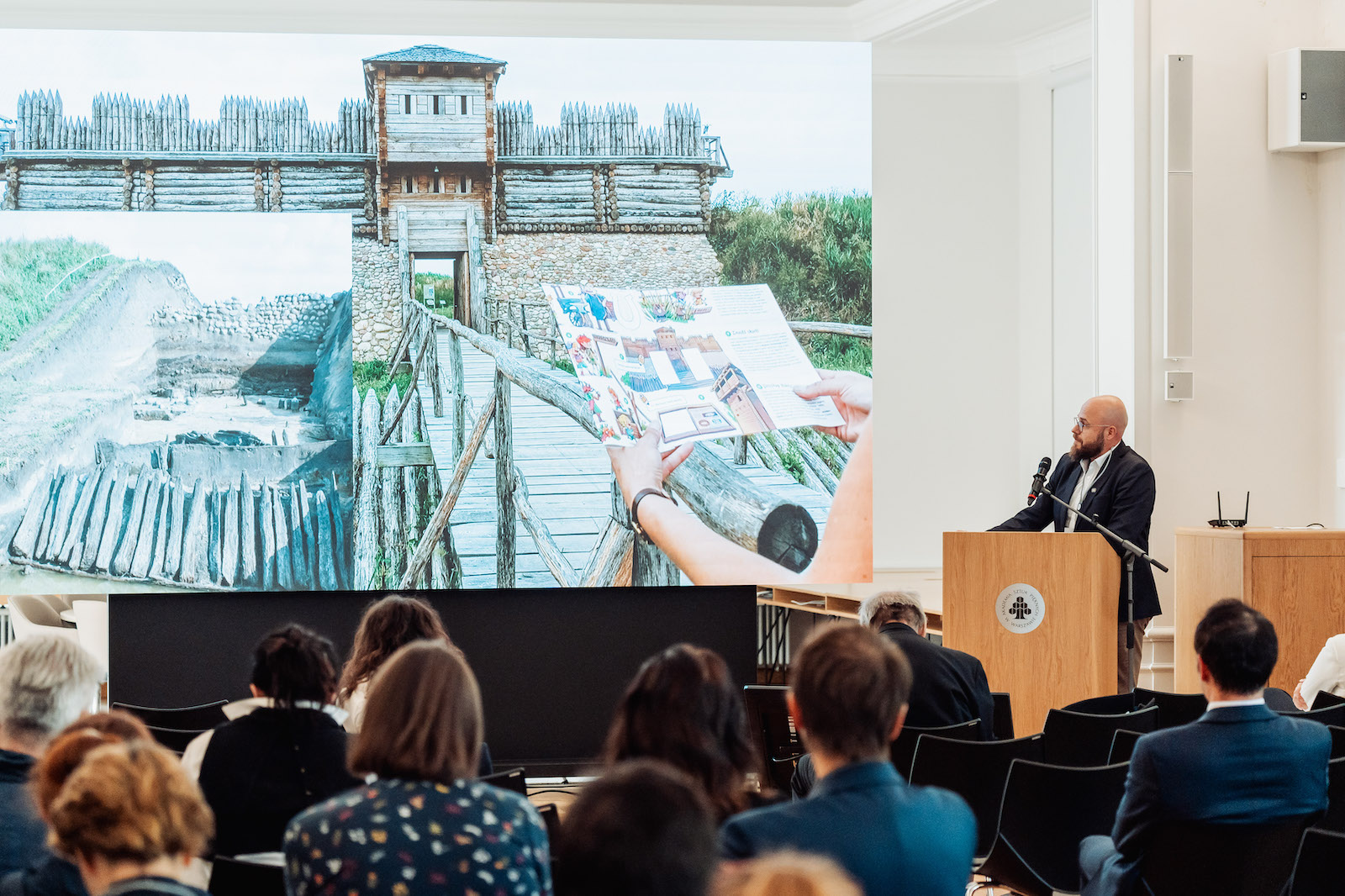 Dominik Kacper Płaza prowadzi prelekcję podczas konferencji Odbudować nieodbudowane. Obok niego ekran ze zdjęciem rekonstrukcji grodziska w Tumie.