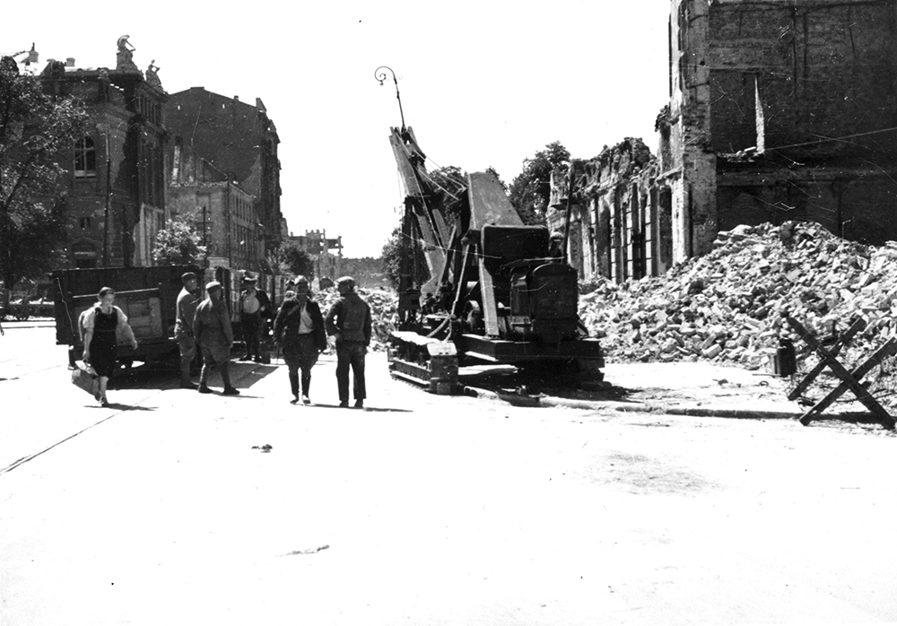Archiwalne zdjęcie zniszczonej ulicy Królewskiej. Po prawej sterty gruzu okalają parterowe pozostałości jednej z kamienic. Na środku stoi dźwig, a obok niego stoi kilka osób.