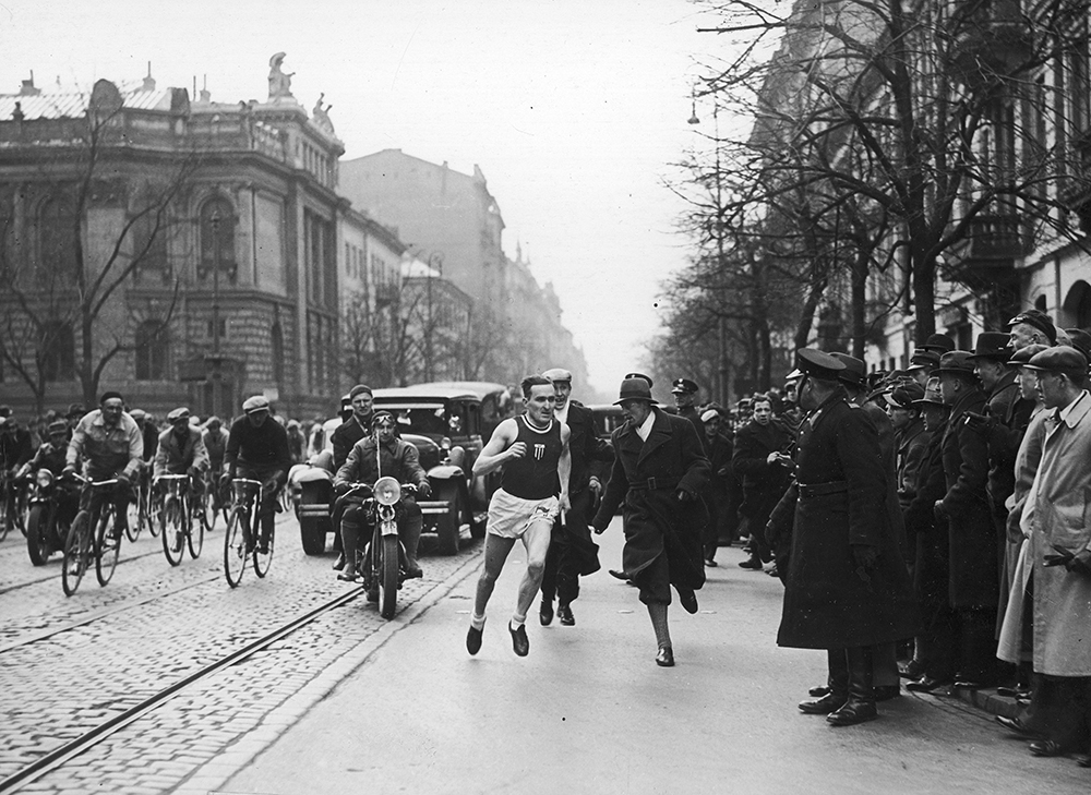 Archiwalne zdjęcie ulicy. Na środku biegnie w krótkim stroju sportowym Janusz Kusociński, za nim jadą ludzie na rowerach i jeden samochód. Z boku, na chodniku stoi tłum gapiów.