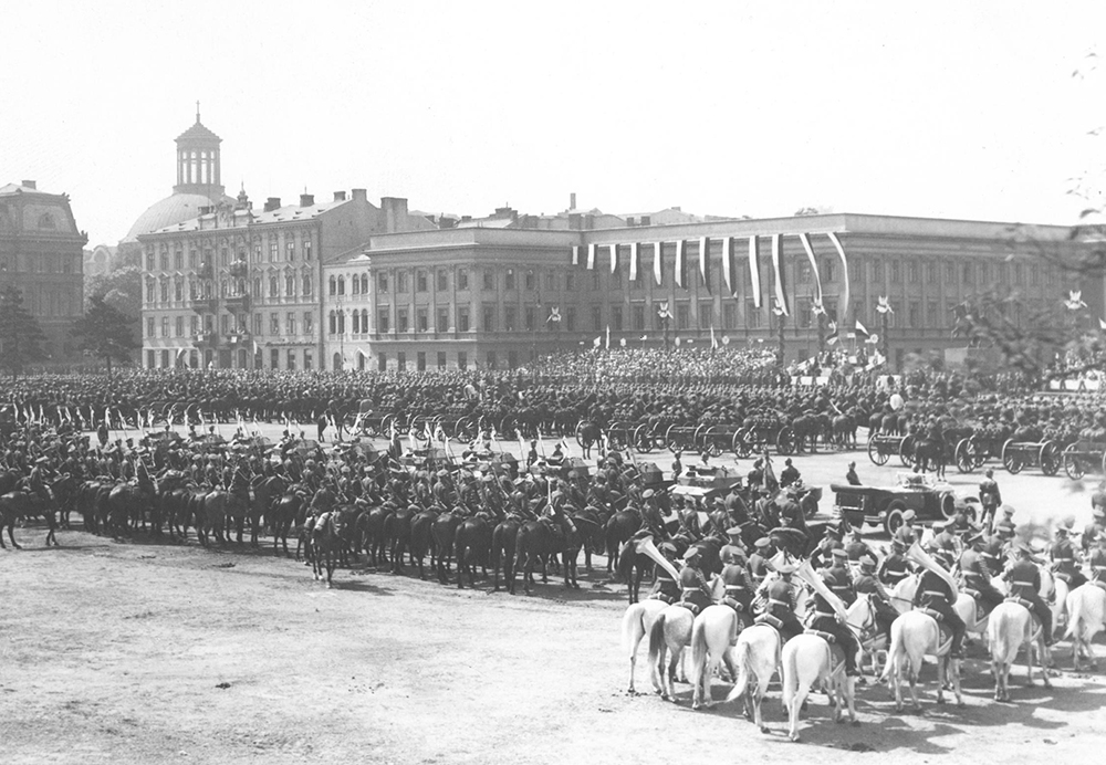 A large square filled with rows of soldiers on horses. In front of them there is the Saski Palace decorated with Polish flags. Next to the palace, there is a corner townhouse with two rows of balconies.