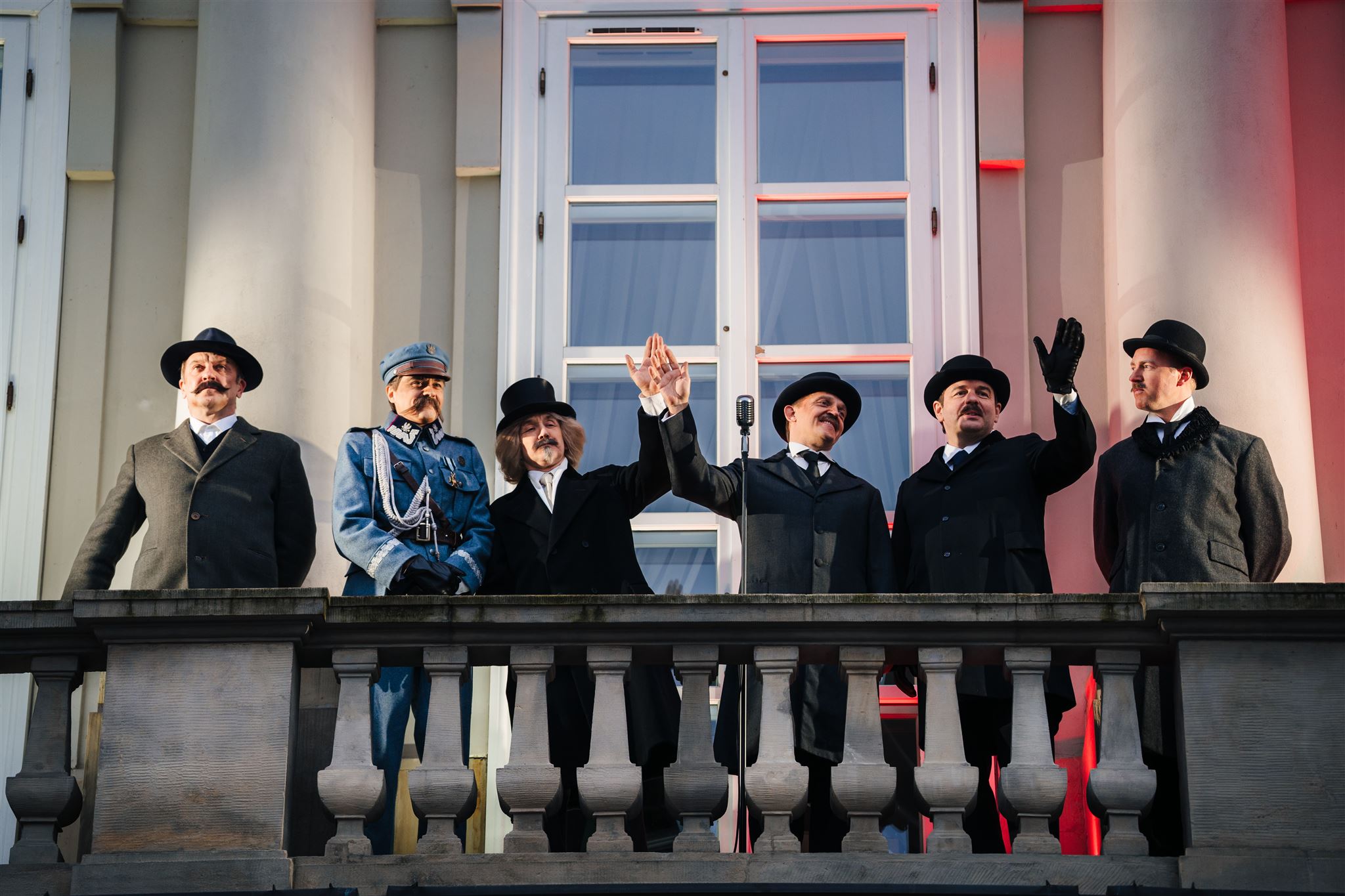 Sześciu mężczyzn w strojach z okresu międzywojennego macha z balkonu. Każdy z nich ucharakteryzowany jest tak, by przypominał jednego z ojców polskiej niepodległości.