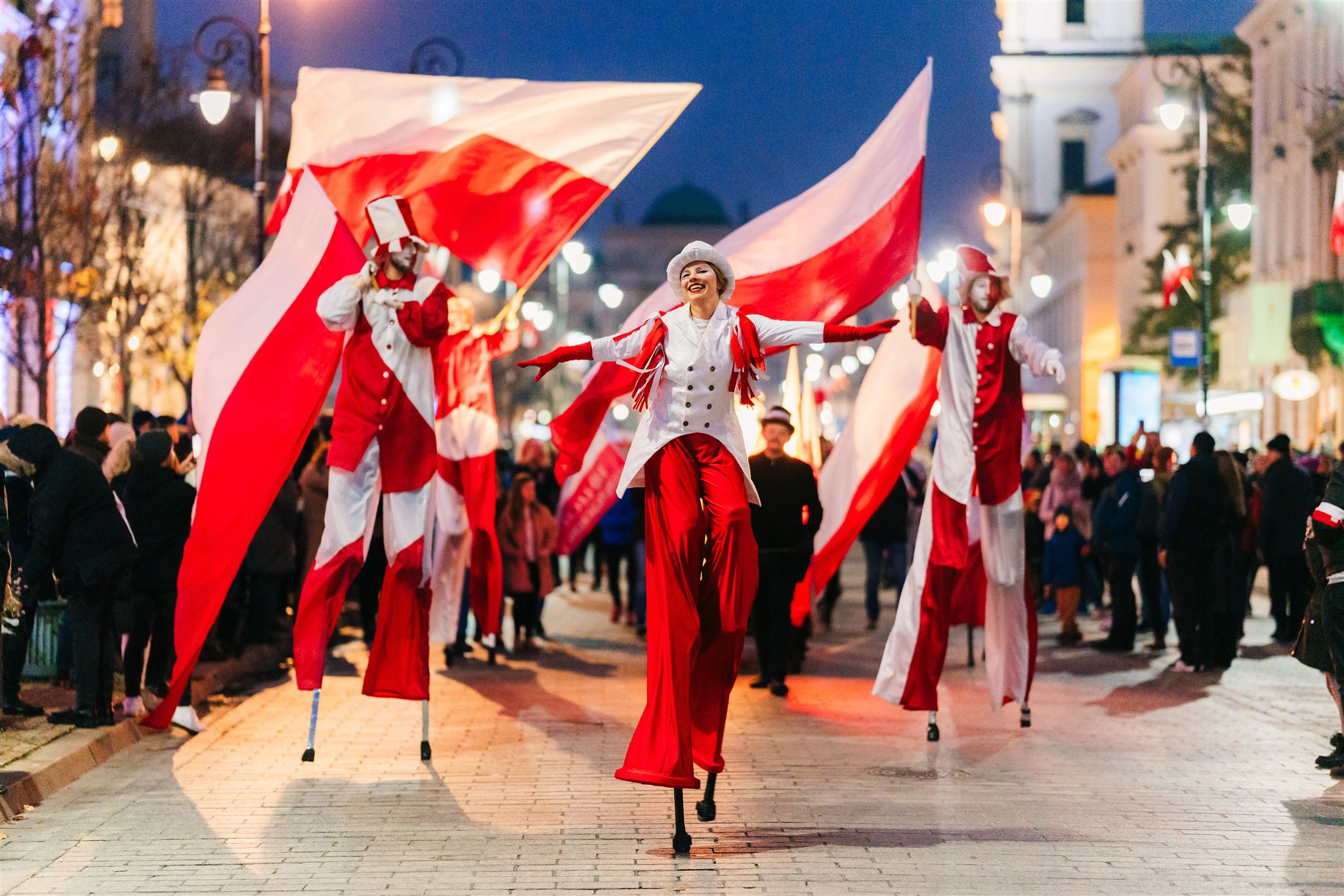 Grupa szczudlarzy krocząca środkiem ulicy podczas Festiwalu Niepodległa na Krakowskim Przedmieściu. Osoby na szczudłach niosą wielkie flagi Polski i są ubrane na biało i czerwono.
