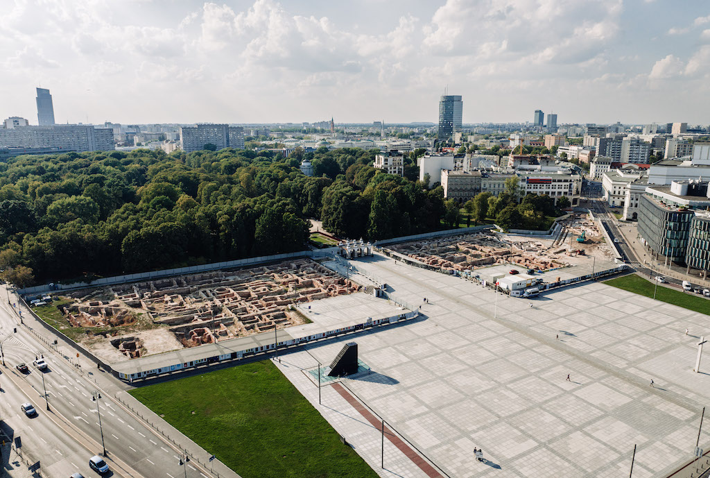 Zdjęcie placu Piłsudskiego wykonane z drona. Część placu jest odgrodzona. Za ogrodzeniem widać odsłonięte, zabytkowe fundamenty. W tle terenu inwestycji bujne drzewa Ogrodu Saskiego i fragment panoramy miasta.