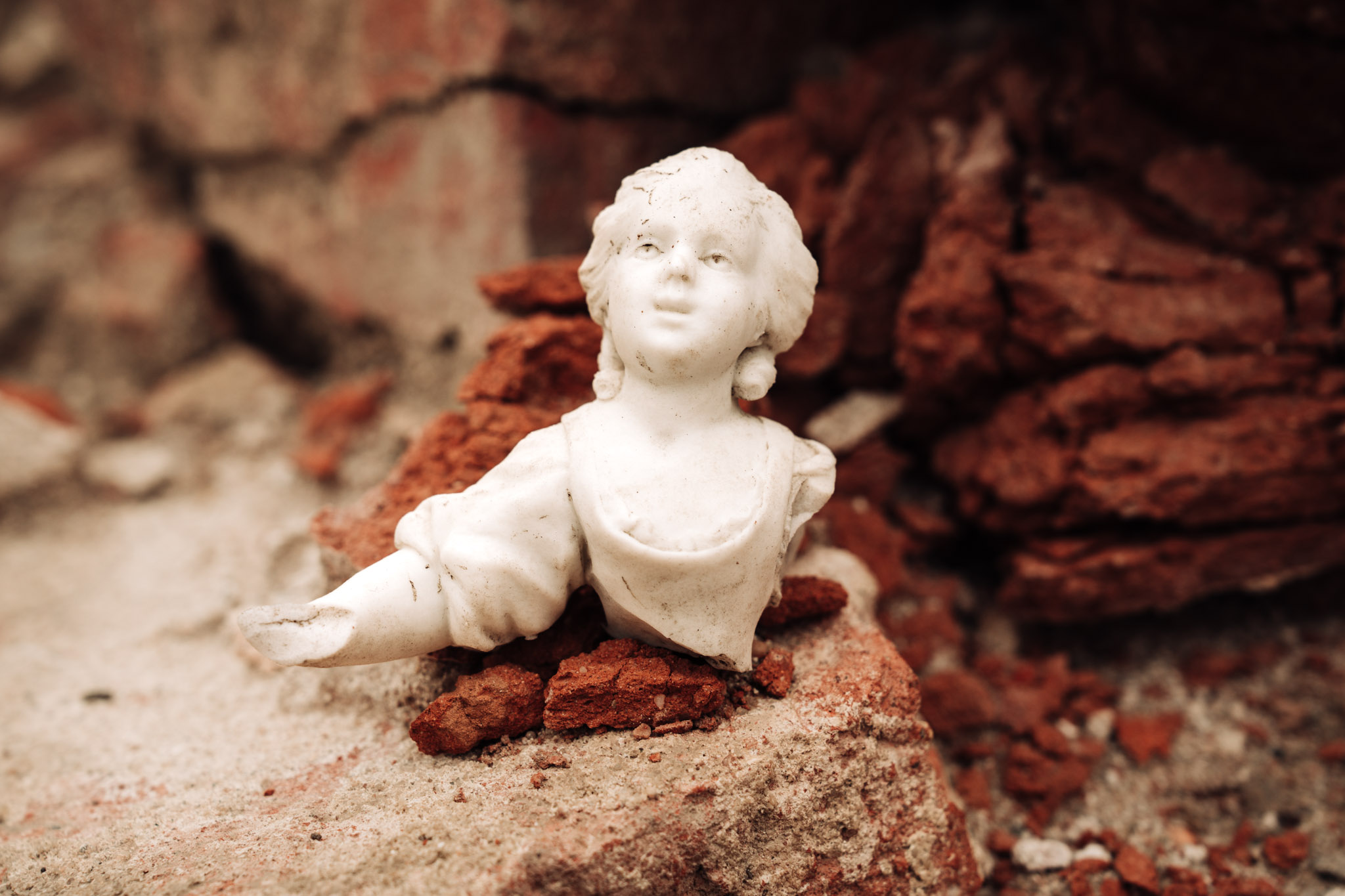 Górna część białej porcelanowej figurki przedstawiającej postać kobiecą. Głowa uniesiona do góry, prawa ręka wyciągnięta do przodu. W tle pozostałości ceglanych murów Pałacu Saskiego.