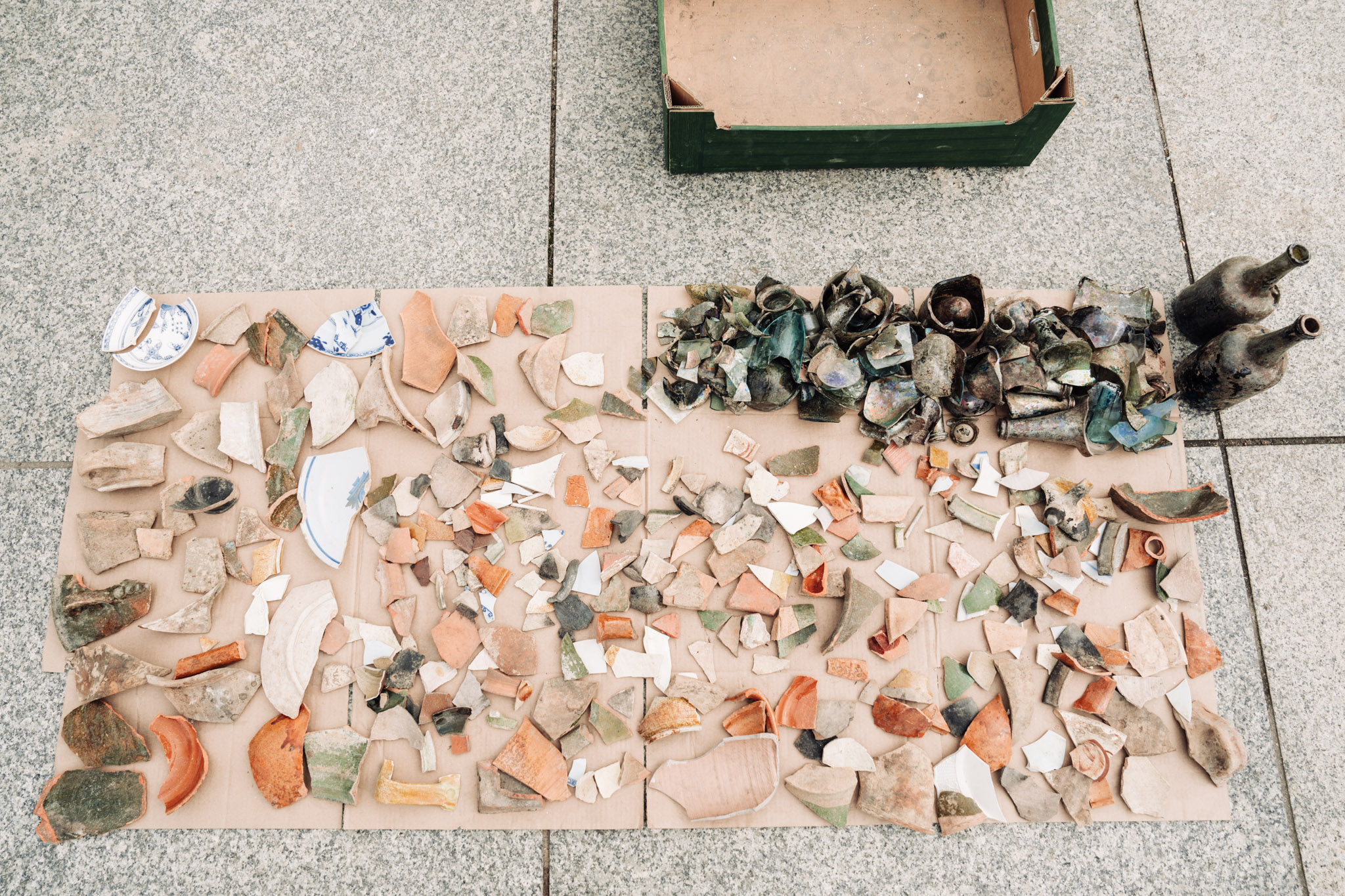 Liczne fragmenty ceramiki i szkła oraz dwie butelki, czyli zagadki archeologiczne czekające na rozwikłanie. Drobne elementy rozłożone są na tekturze znajdującej się na granitowych płytach.