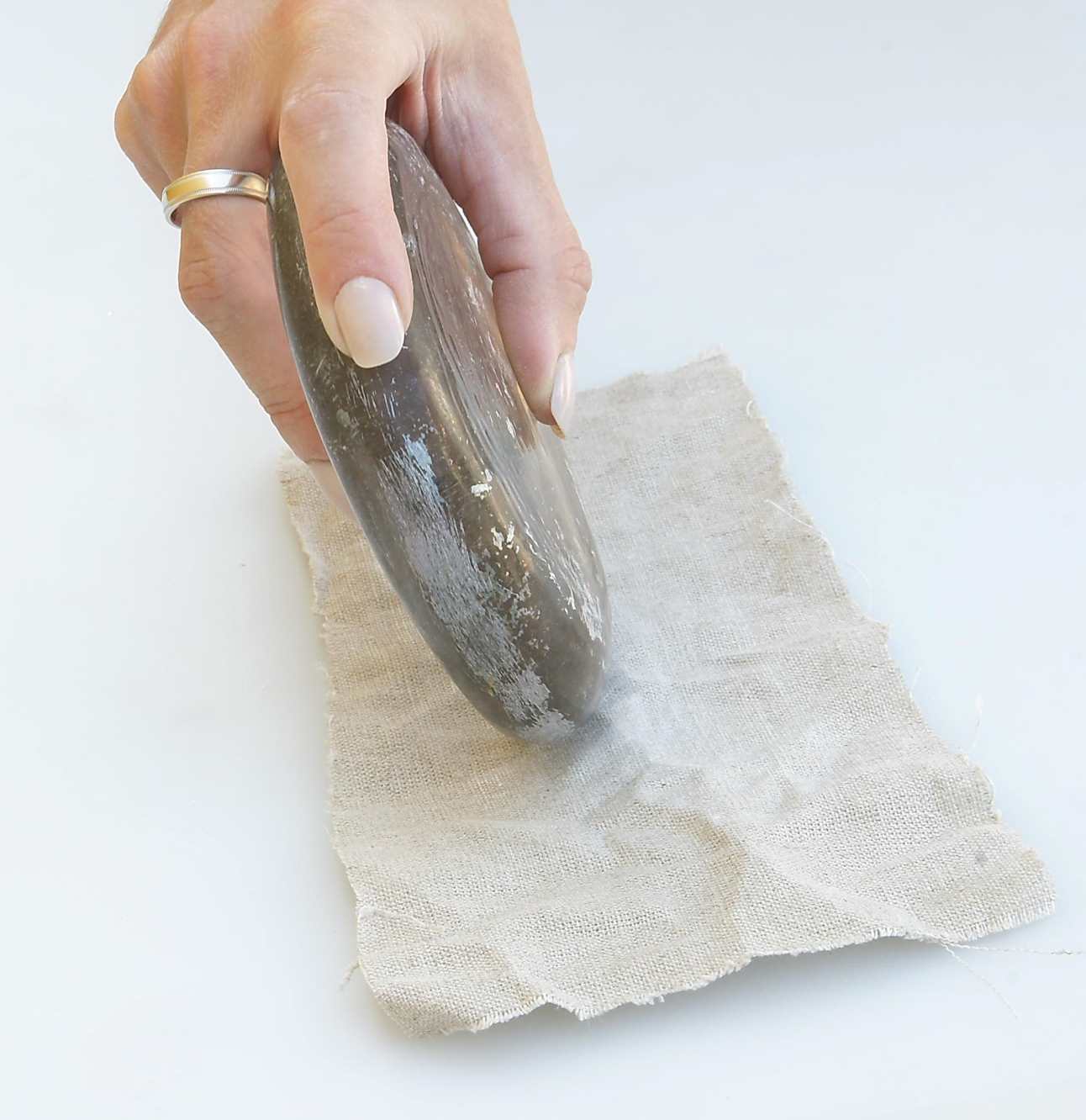 Szklany, okrągły przedmiot w kobiecej dłoni to jeden z egzemplarzy zagadki archeologicznej. Na zdjęciu użyty do prasowania kawałka lnu. Białe tło.