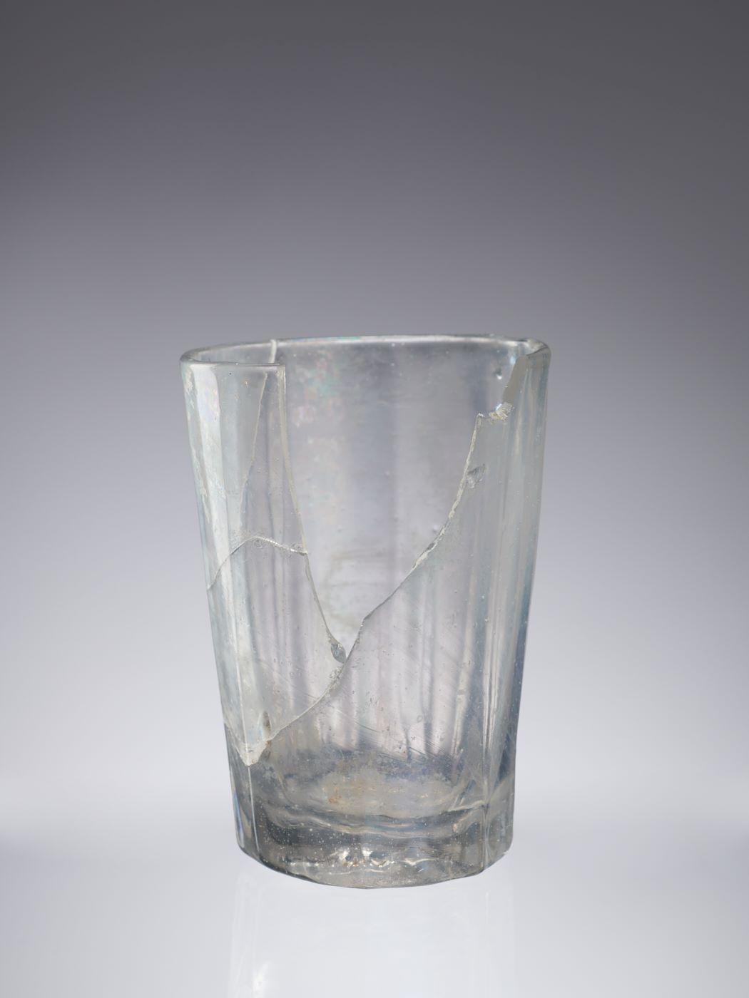 Popękana i pozbawiona fragmentu szkła w górnej części, przezroczysta szklanka, zwężona u dołu, z grubym dnem.