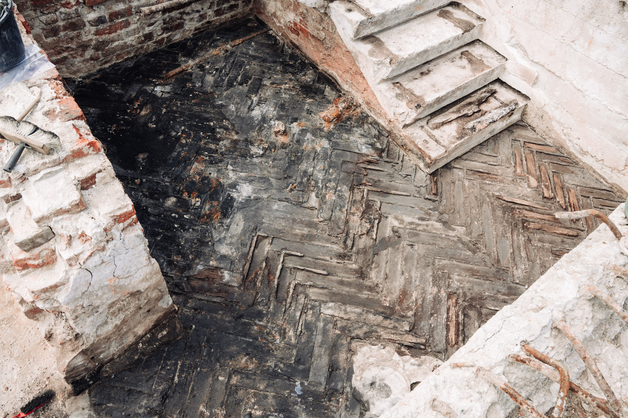 Zdjęcie pokazuje badania archeologiczne Pałacu Brühla. Podłoga pomieszczenia, w której odciśnięte są wgłębienia po podłużnych klepkach ułożonych w jodełkę. W jednym z rogów pomieszczenia znajdują się uszkodzone schody.