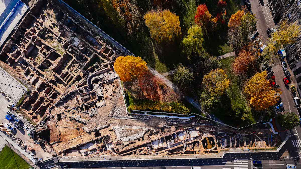Fotografia z drona przedstawiająca z góry widok na badania archeologiczne na terenie Pałacu Brühla, czyli plątaninę murowanych fundamentów odsłoniętych w ziemi. Obok wykopalisk z jednej strony prowadzi jezdnia, z drugiej znajdują się drzewa i trawa.