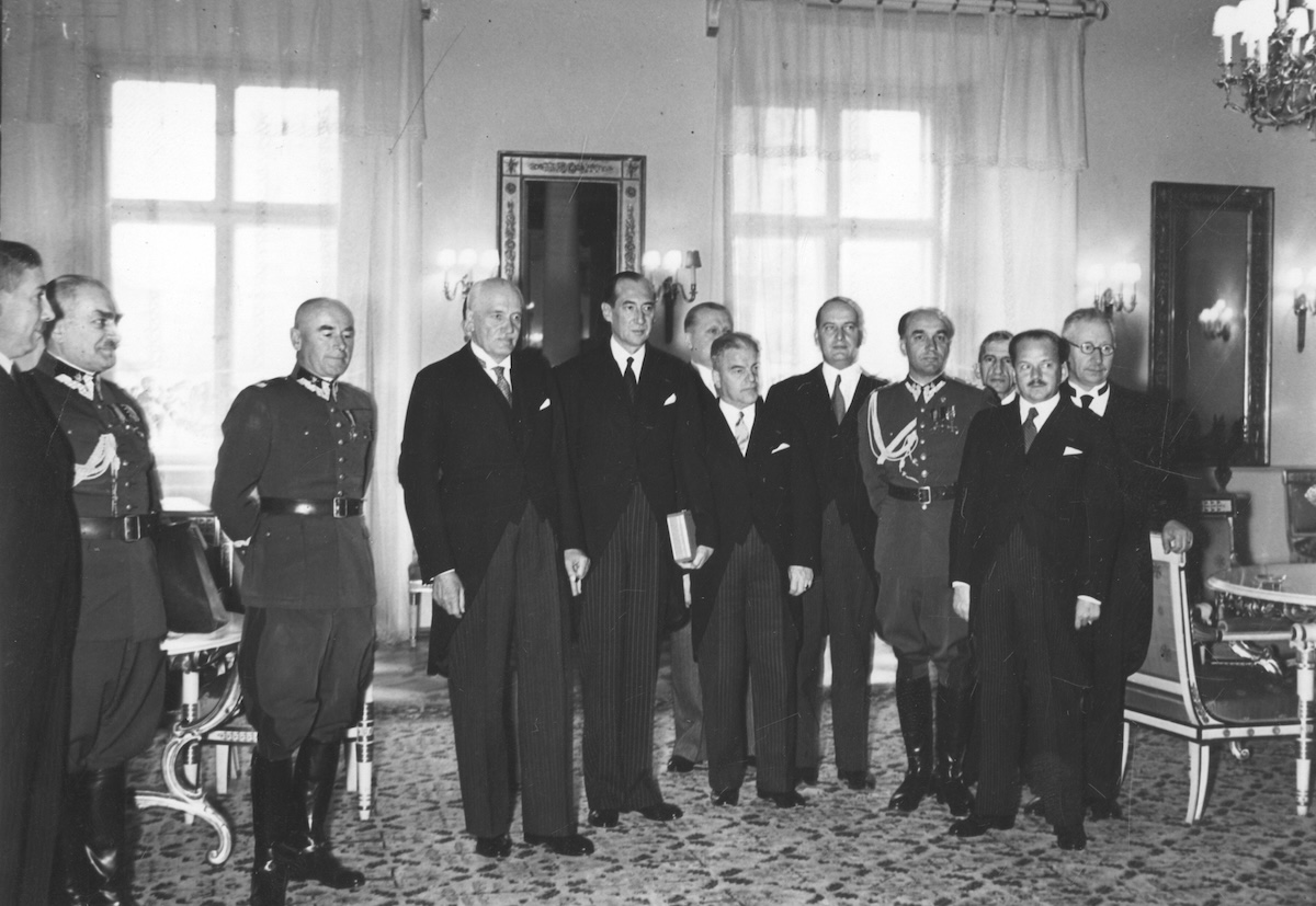 Czarno-biała fotografia, na której stoi szereg mężczyzn w bogato zdobionym wnętrzu. Część ubrana jest we fraki, część w mundury Wojska Polskiego.