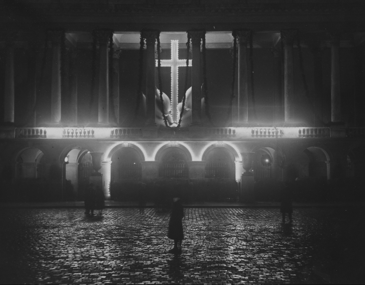 Podświetlona kolumnada Pałacu Saskiego wyłaniająca się z ciemności nocy. Po środku podświetlony jest Grób Nieznanego Żołnierza, a nad jego arkadami świecący krzyż pomiędzy kolumnami.