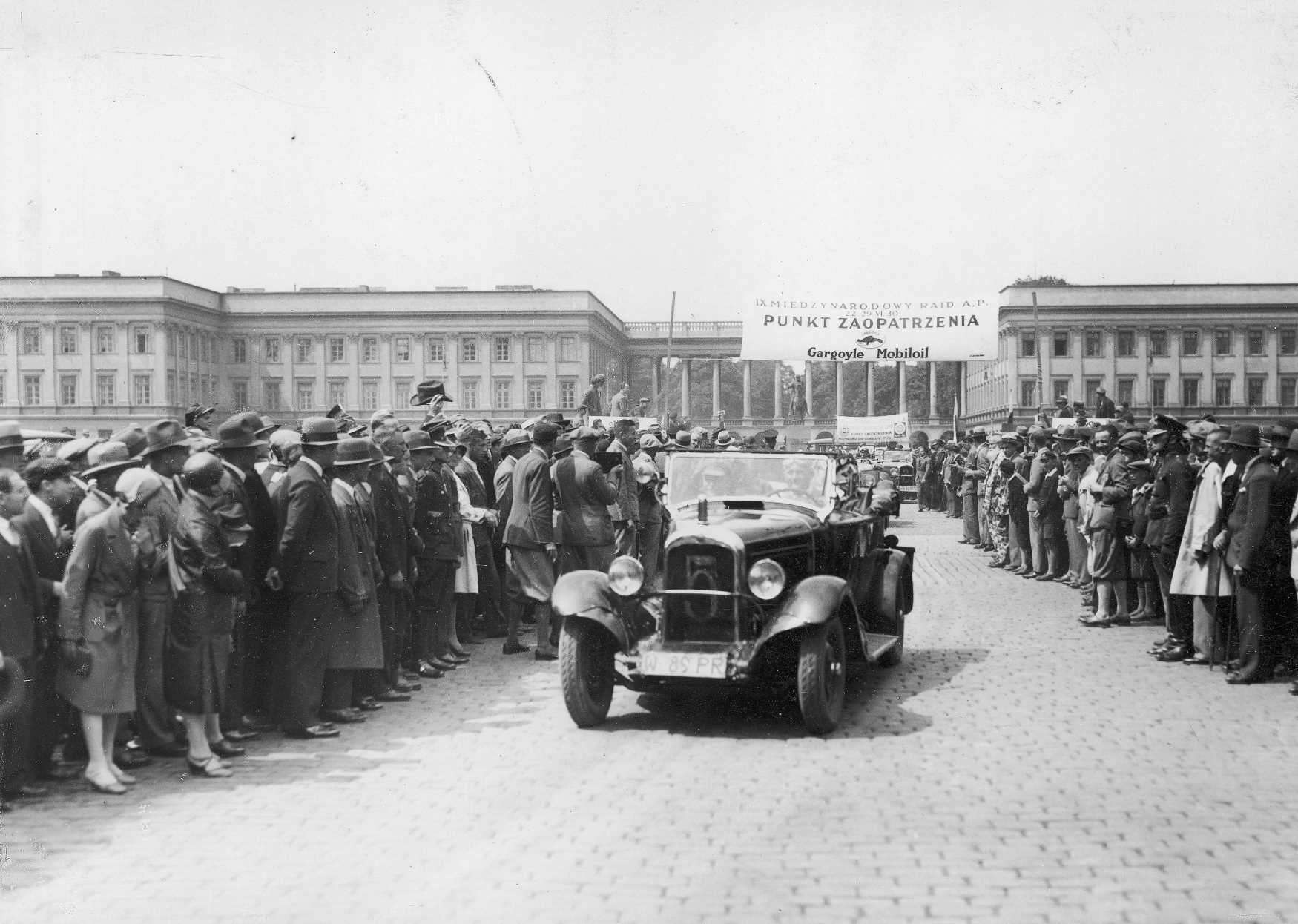 Plac Piłsudskiego, czyli dawny plac Saski, zdjęcie z okresu międzywojennego. Środkiem jedzie kabriolet, po obu stronach stoją tłumy gapiów. W tle wyłania się bryła Pałacu Saskiego.