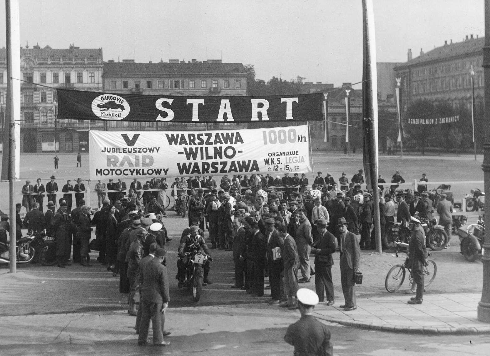 Plac Piłsudskiego, czyli dawny plac Saski, zdjęcie z okresu międzywojennego. Między dwoma masztami flagowymi rozwieszone są dwa pasy z napisami start i nazwą rajdu motocyklowego Warszawa-Wilno-Warszawa. Pod nimi wyjeżdża motocyklista otoczonymi gapiami. W tle pierzeje kamienic okalające plac.