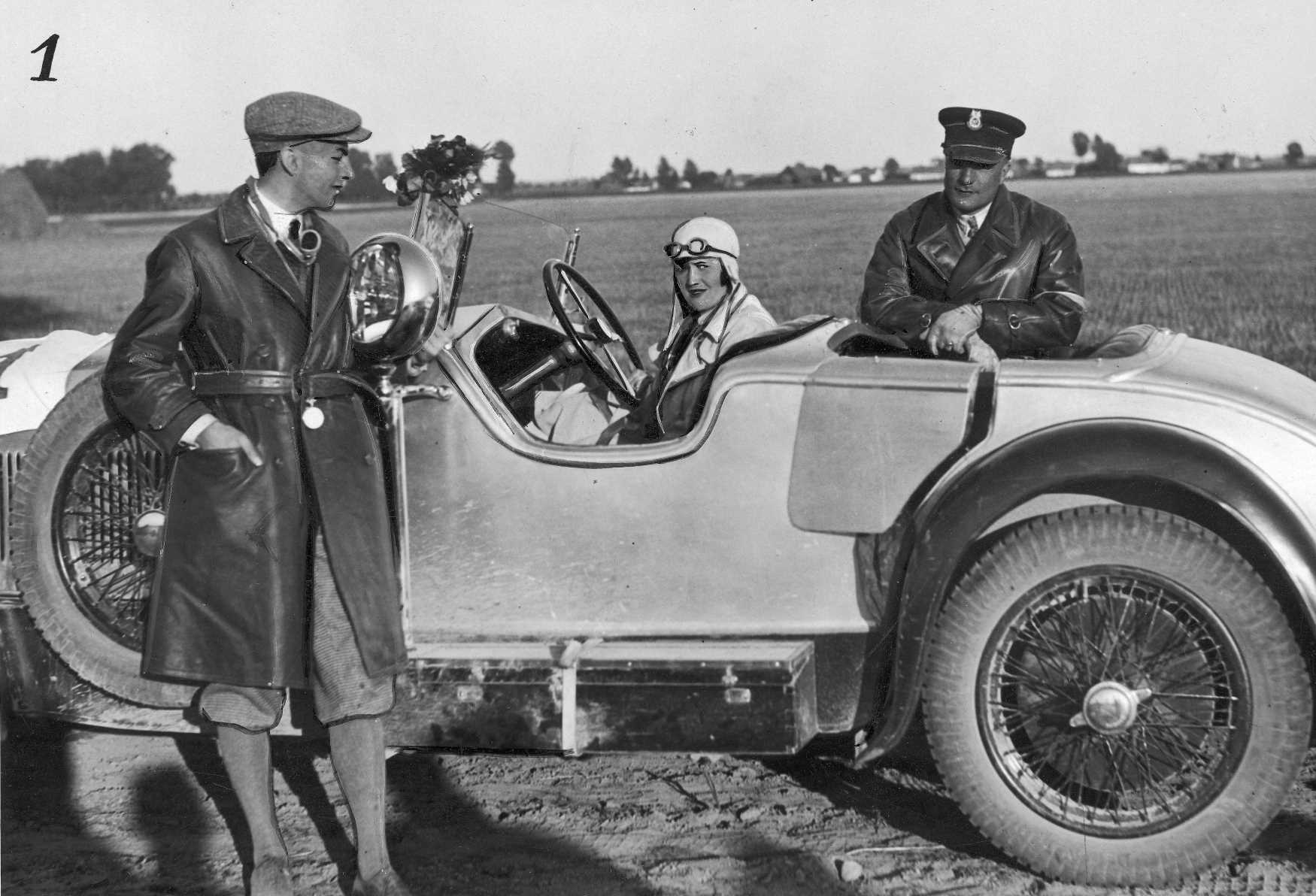 Przedwojenne zdjęcie. Kobieta siedzi w sportowym kabriolecie. Po obu stronach samochodu stoją mężczyźni w skórzanych płaszczach z kołnierzami. W tle, za samochodem, znajduje się pole.