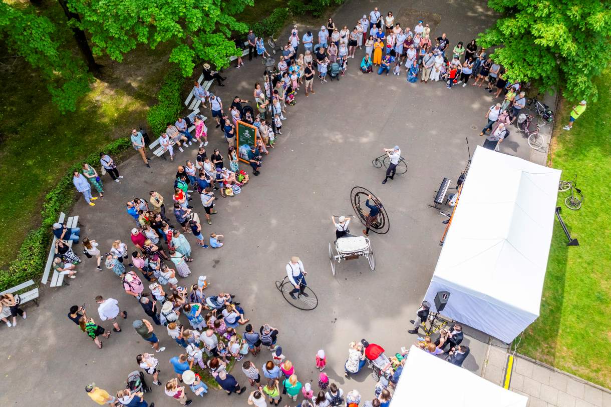 Relacja ze Święta Ogrodu Saskiego z drona. Widok z góry na pokaz dawnych bicykli przed namiotem w alei Ogrodu Saskiego. Dookoła ludzi na rowerach tłum gapiów.