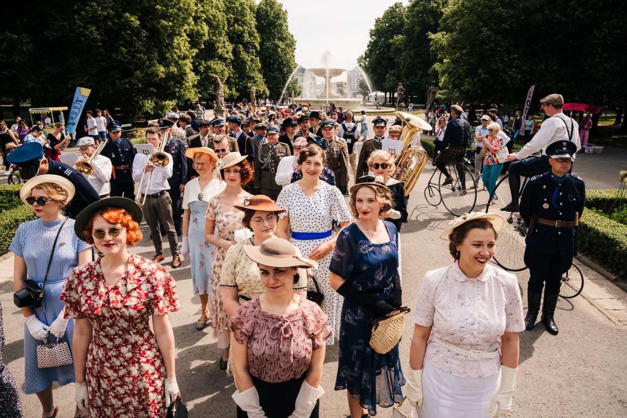 Tłum ludzi w alei głównej Ogrodu Saskiego. Na pierwszym planie kobiety w sukienkach i kapeluszach z okresu międzywojennego, za nimi zespół muzyczny z trąbkami i puzonami. W głębi mężczyźni w mundurach wojskowych i eleganckich ubraniach z tej samej epoki. Obok dwóch mężczyzn w kaszkietach jeździ na rowerach z wielkim przednim kołem i małym tylnym. W tle fontanna. 