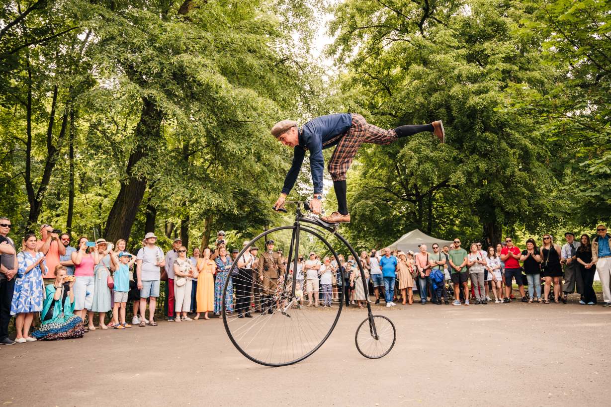 Mężczyzna w kaszkiecie i pumpach w kratę stoi na jednej nodze na siodełku roweru. Rower ma wielkie przednie koło i maleńkie tylne. Dookoła tłum gapiów w różnym wieku, za nimi drzewa Ogrodu Saskiego.