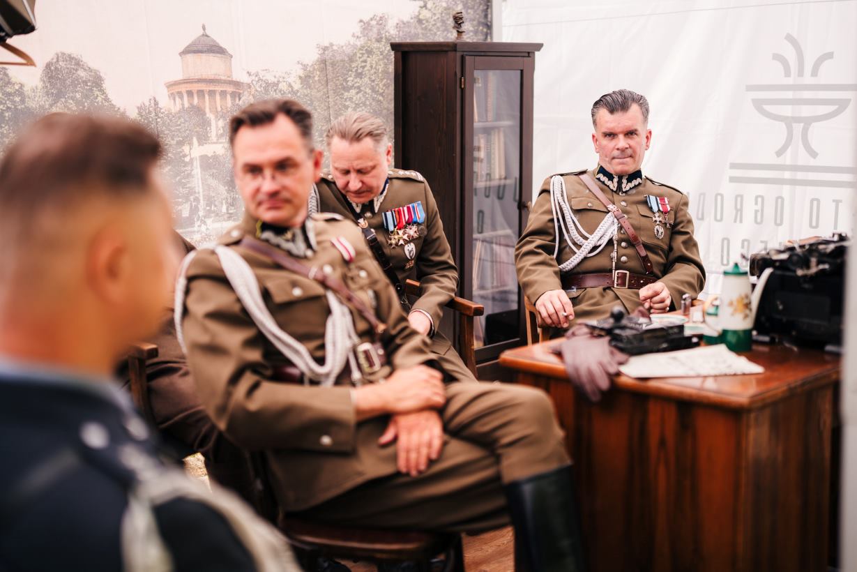 Trzech mężczyzn w polskich mundurach wojskowych z okresu międzywojnia siedzi wokół drewnianego biurka.