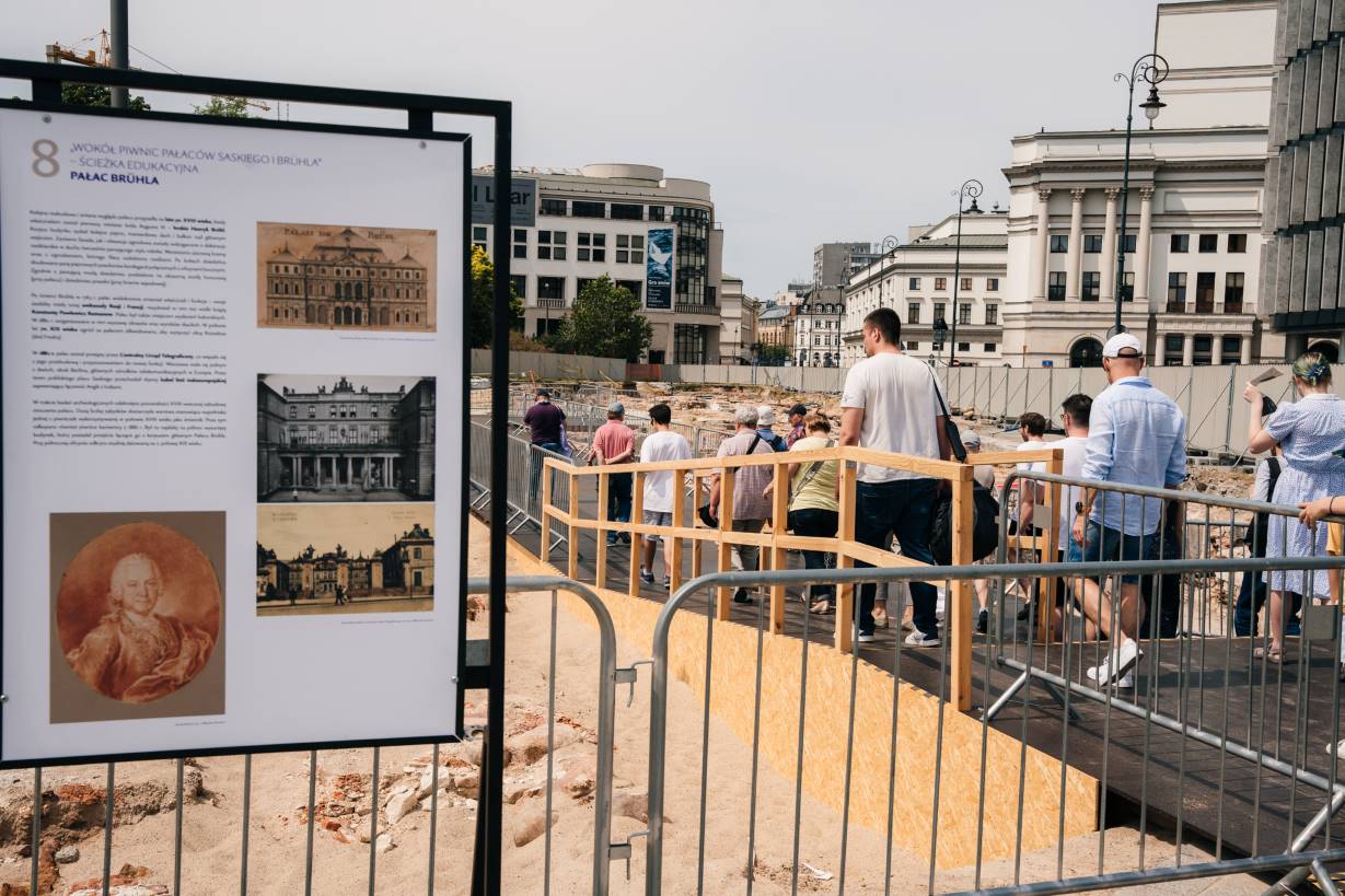 Grupka osób idzie podestem ustawionym nad odsłoniętymi fundamentami. Po bokach ludzi barierki zabezpieczają trasę. Na pierwszym planie tablica edukacyjna z opisem oraz ilustracjami pokazującymi jak kiedyś wyglądał Pałac Bruhla i sam właściciel, Henryk Bruhl.