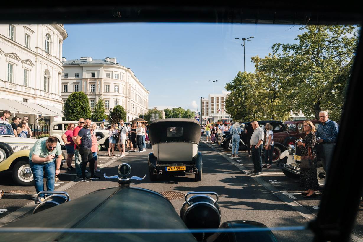 Widok z miejsca pasażera w starym automobilu na jadące przed nim przedwojenne auto. Po bokach zaparkowane inne stare wozy, pomiędzy którymi stoją tłumy podziwiające auta.