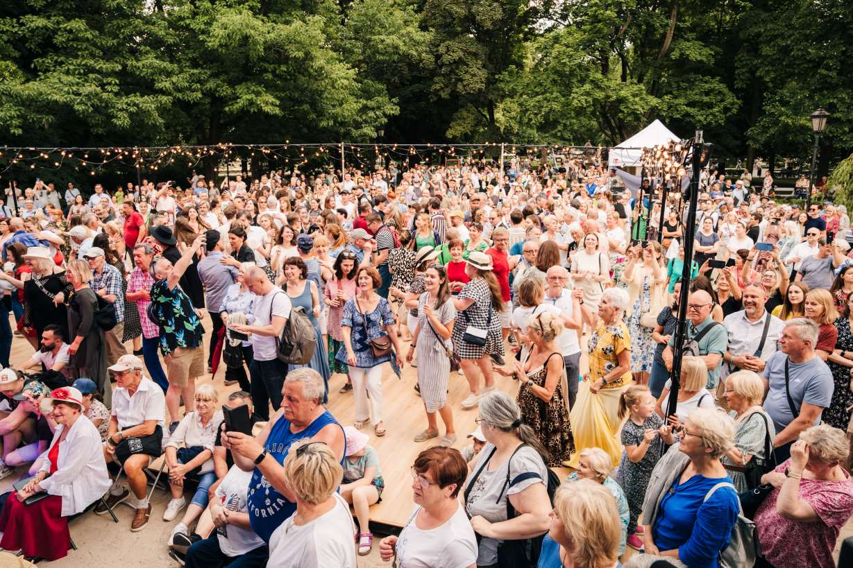 Tłumy ludzi stoją na drewnianym parkiecie pośród drzew.