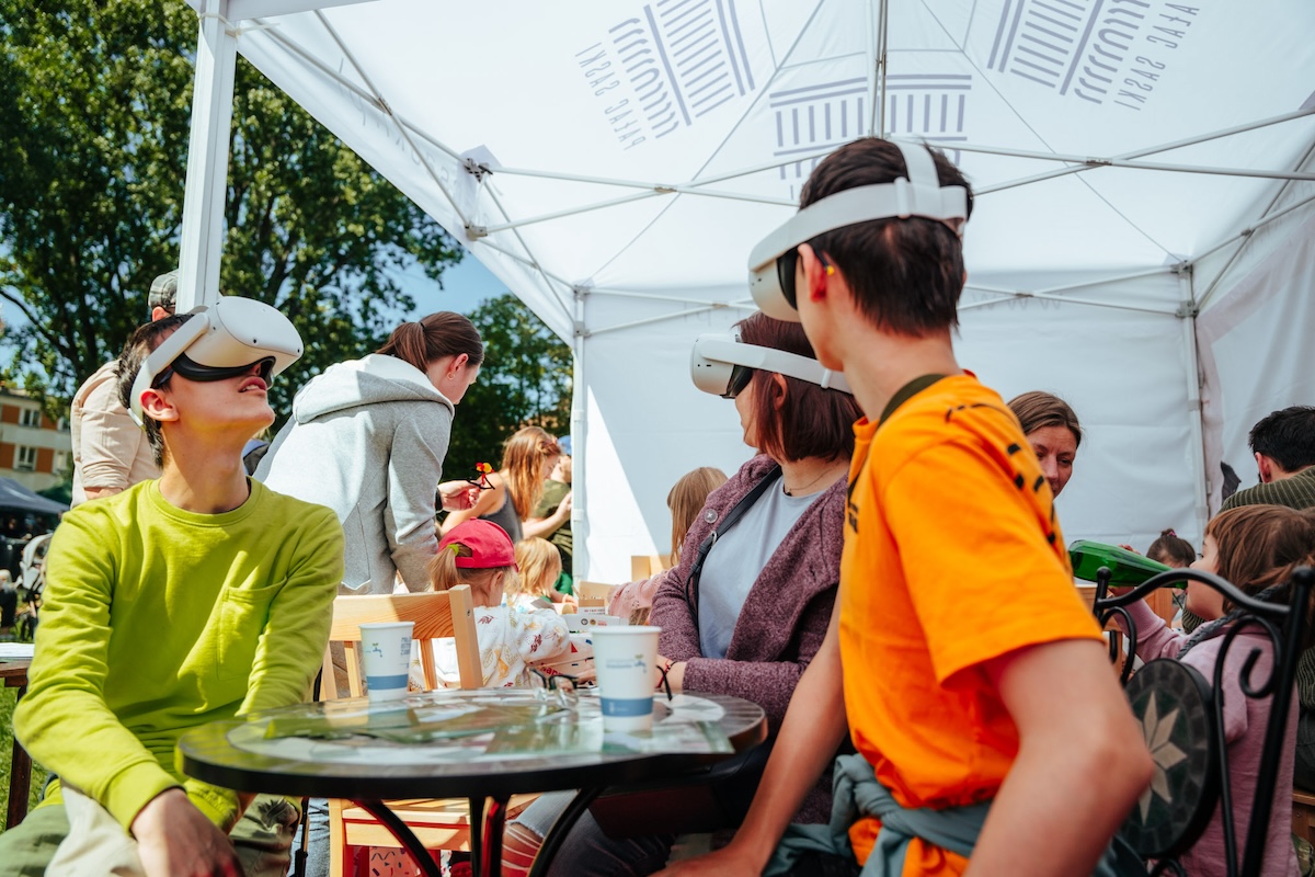 Stoisko Pałac Saski po sąsiedzku w plenerze. Pod namiotowym zadaszeniem siedzi trójka osób przy retro stoliczku. Każda z osób ma na głowie założone gogle VR, które zasłaniają połowę twarzy. Za nimi dzieci z nadzorem dorosłych bawią się przy stole.