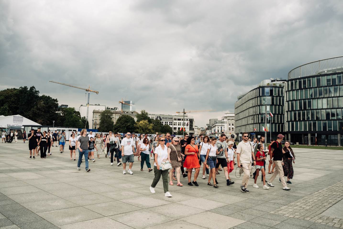 Grupa ludzi idąca przez plac marszałka Józefa Piłsudskiego. W tle zadaszenie reliktów Pałacu Saskiego z logotypem Pałac Saski sp. z o.o. Na dalszym planie drzewa Ogrodu Saskiego oraz budynki i żurawie budowlane. Po prawej stronie budynek Metropolitan.